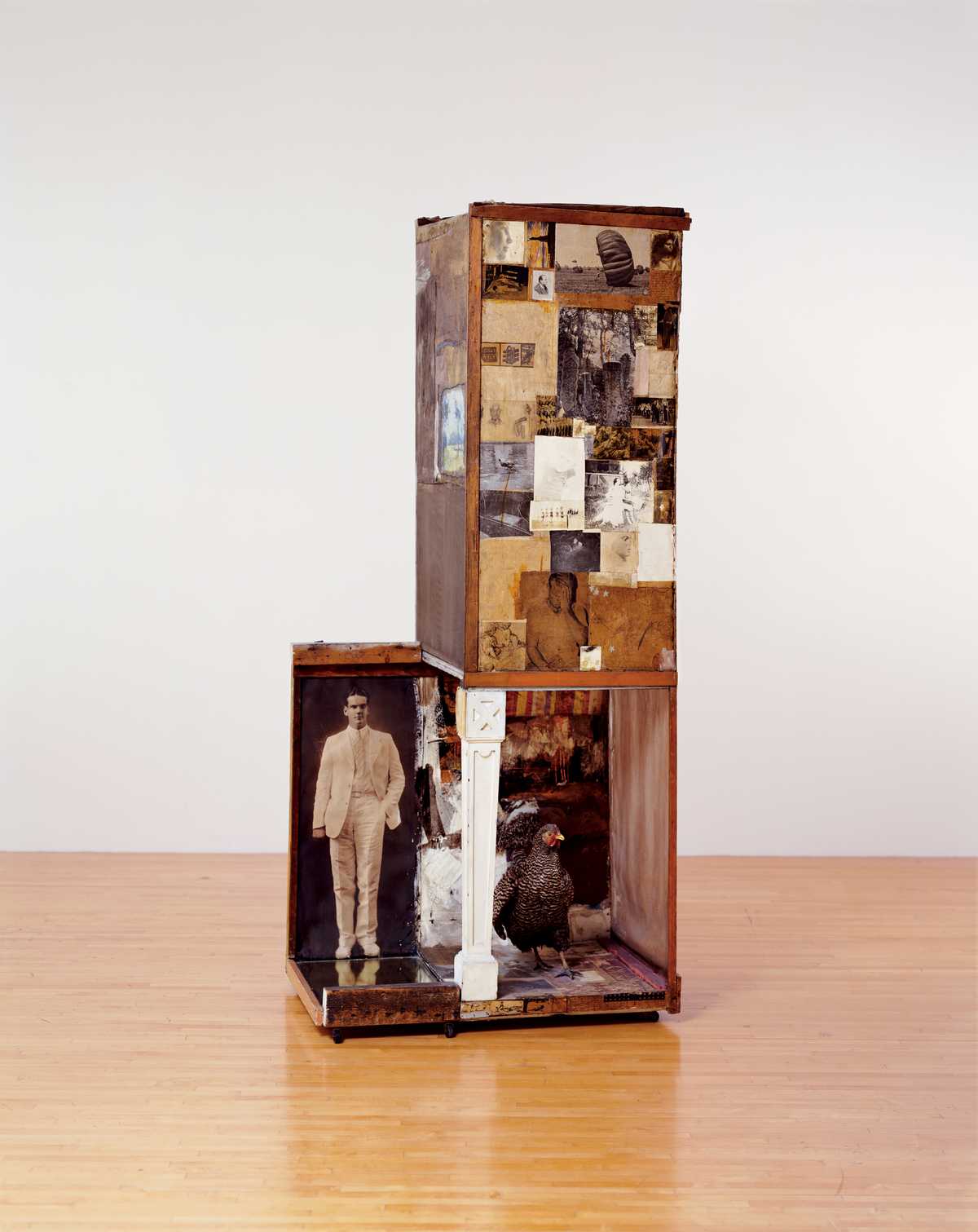 A tall slender wooden box plastered in postcards and new paper clippings, one corner of the box opens and is supported by a white wooden table leg