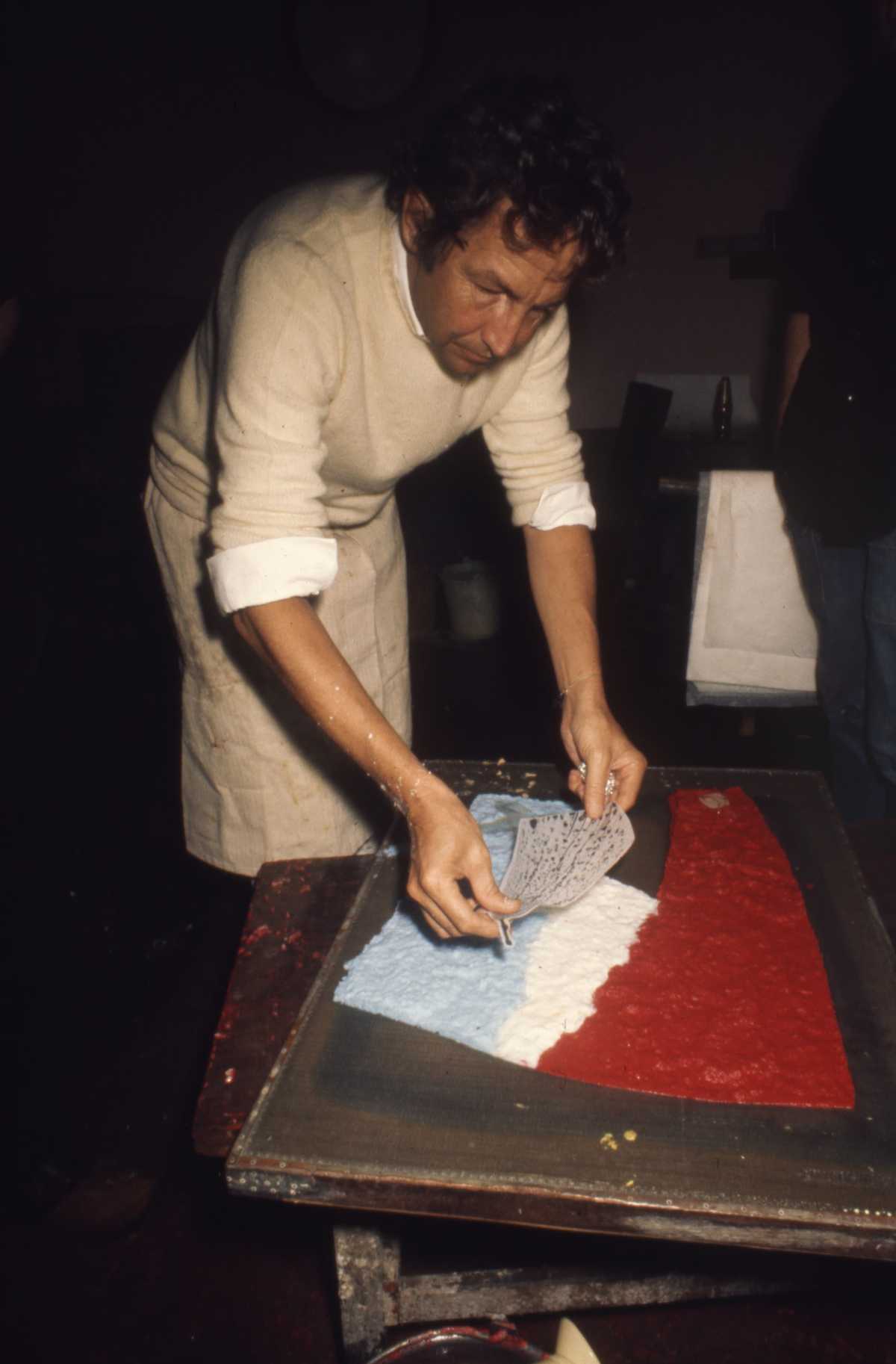 A man leans over a table placing paper over a half complete work of art on the table.