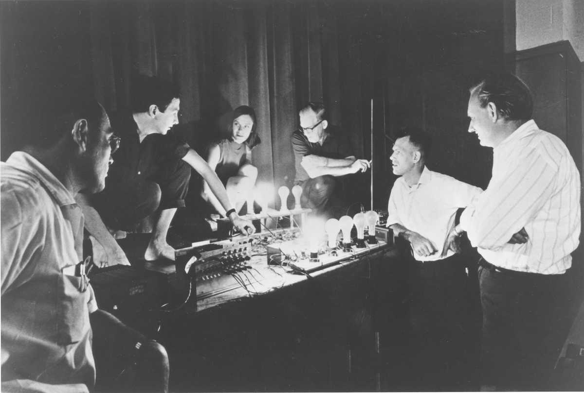 A group of people stand in a darkened room around a collection of lightbulbs on a table