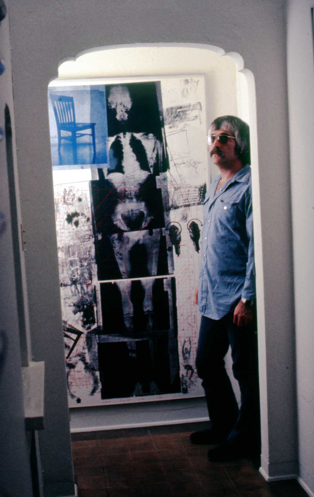 A photo of a man standing in an archway with the print artwork Booster displayed on the wall next to him