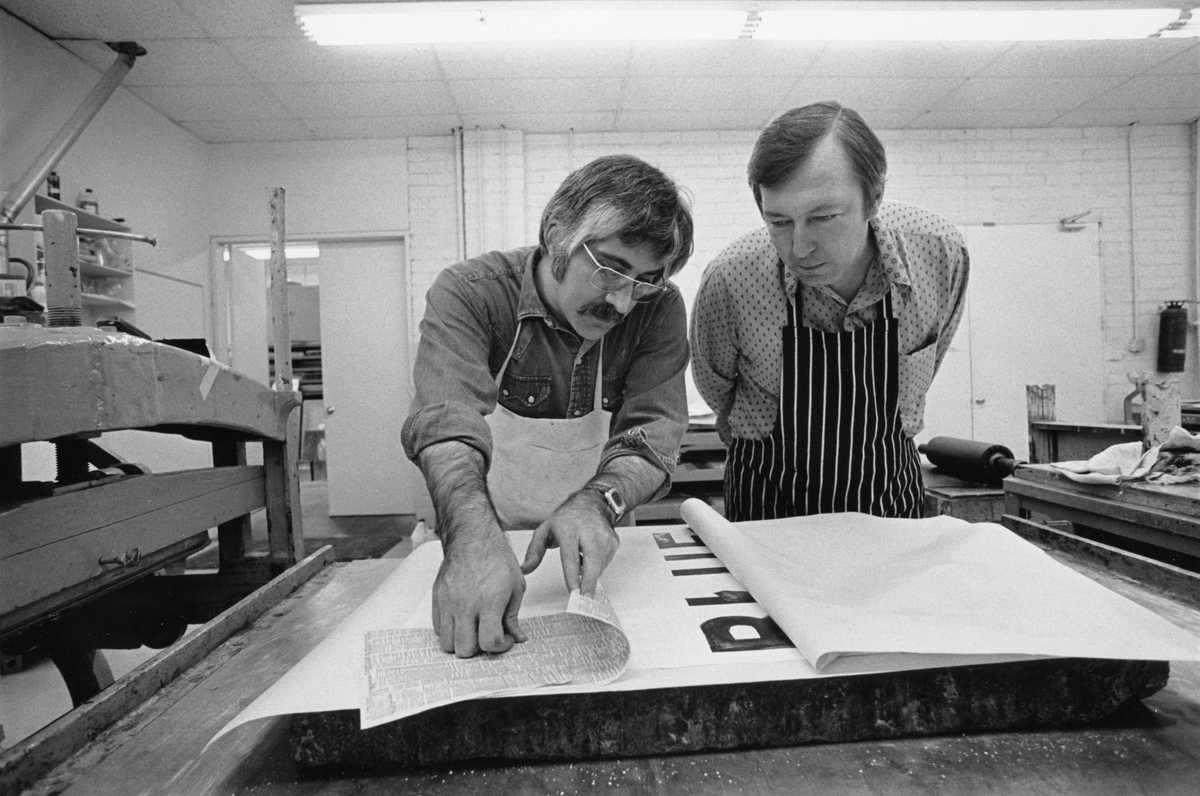 Two men lean over a table towards the camera, both are focused on a print on the table in front of them