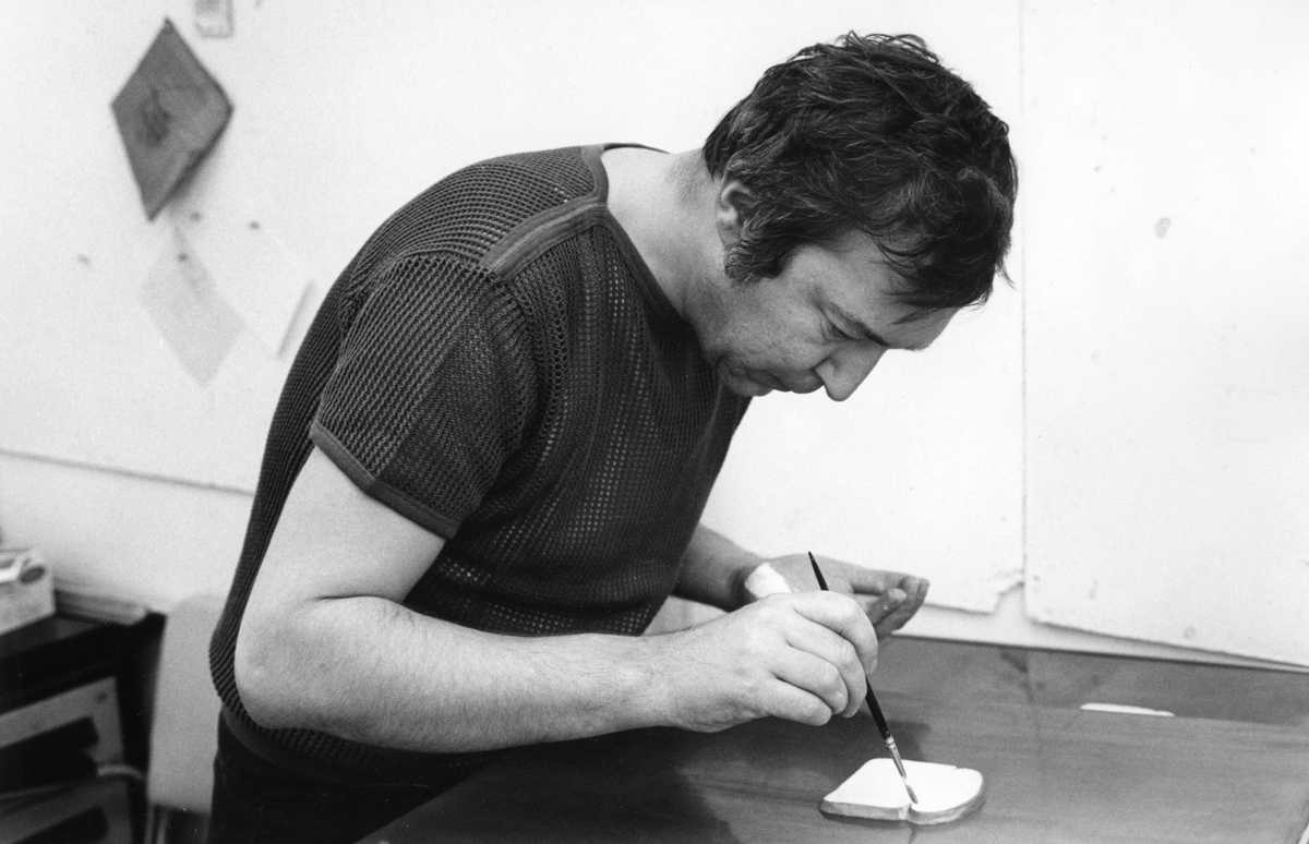 A man sits hunched over a table with a paintbrush in hand, seemingly painting directly onto a piece of bread