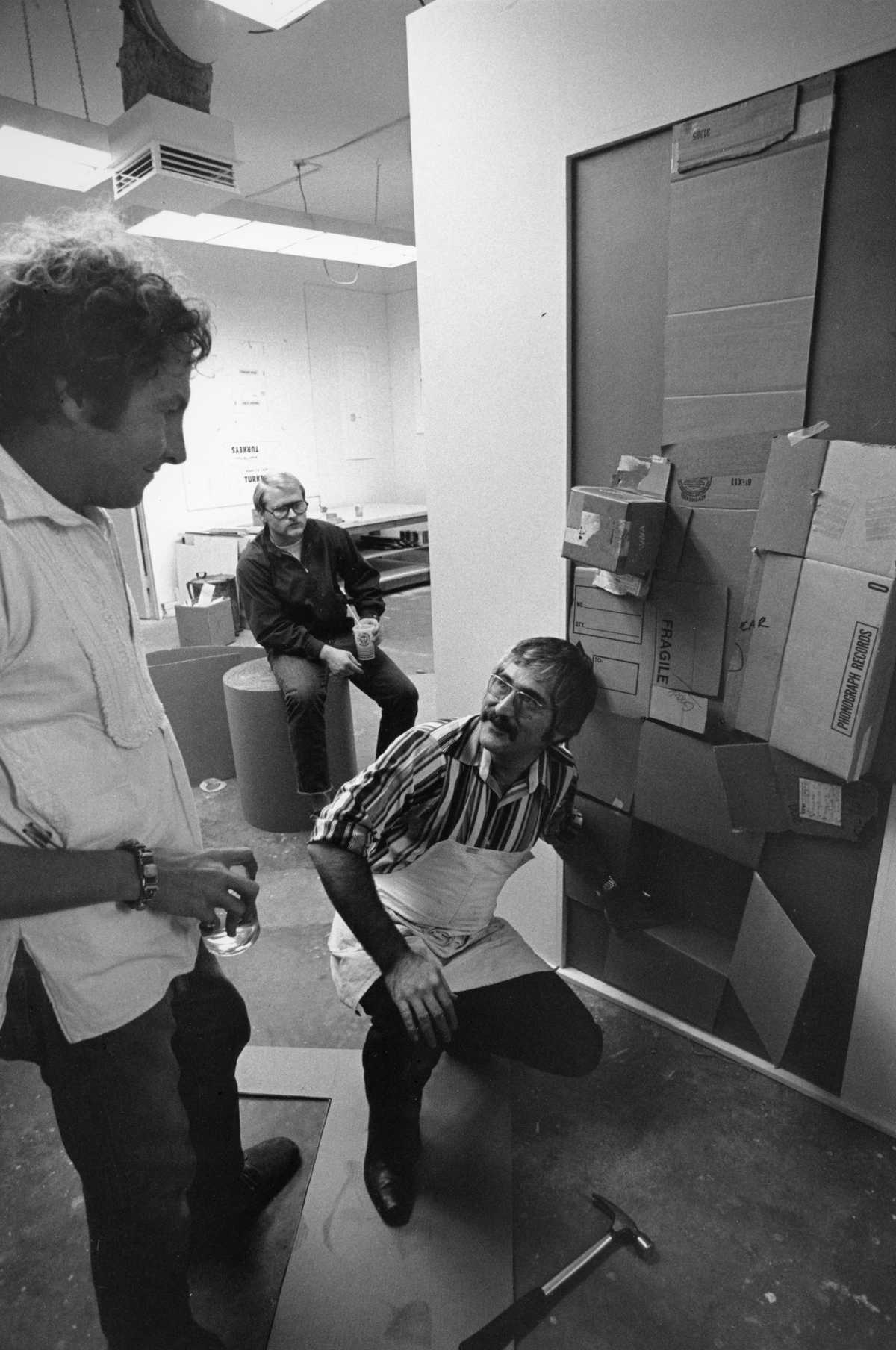 One men crouches on the floor in front of an artwork hung on a wall, talking to a man standing up behind him