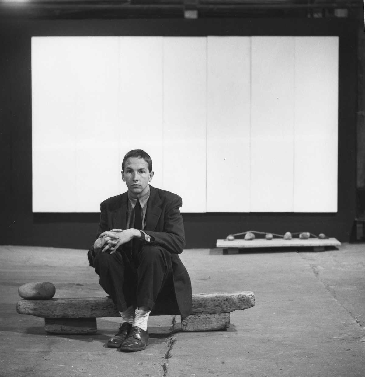 Robert Rauschenberg sits on a log on the floor with "White Painting [seven panel]" in the background