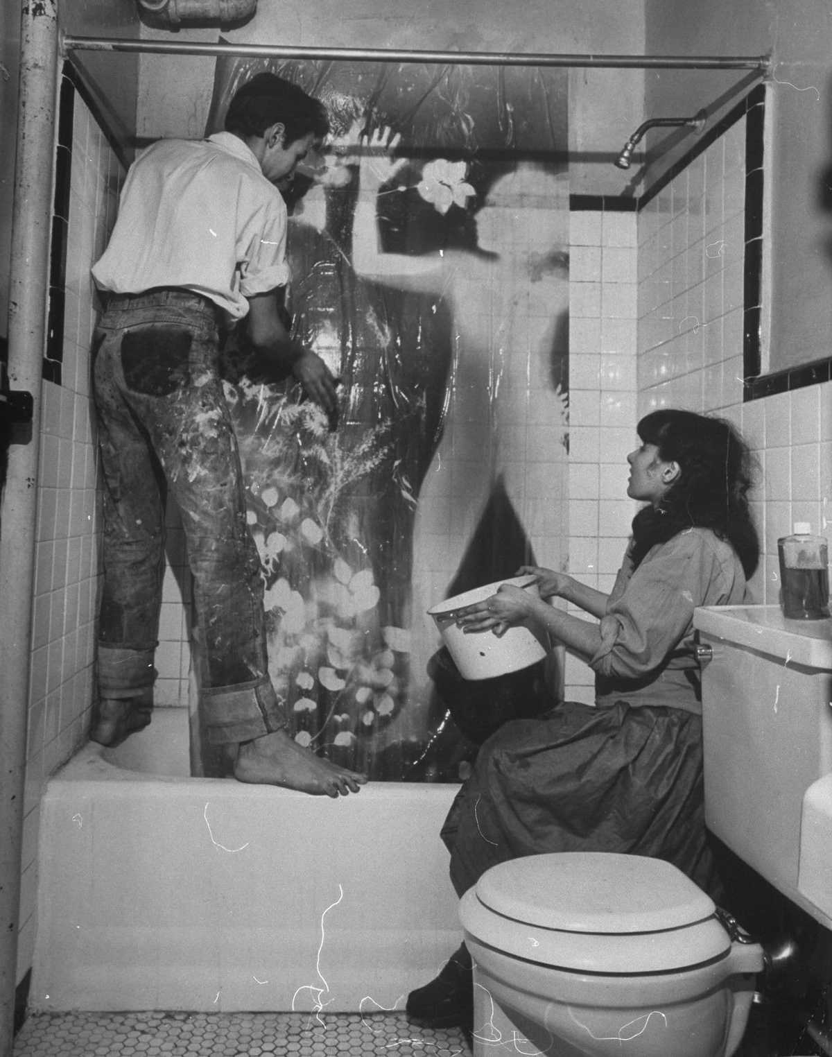 A man stands on a bathtub with an artwork pinned to the wall above the tub. A woman crouches on the floor next to the tub passing something to the man