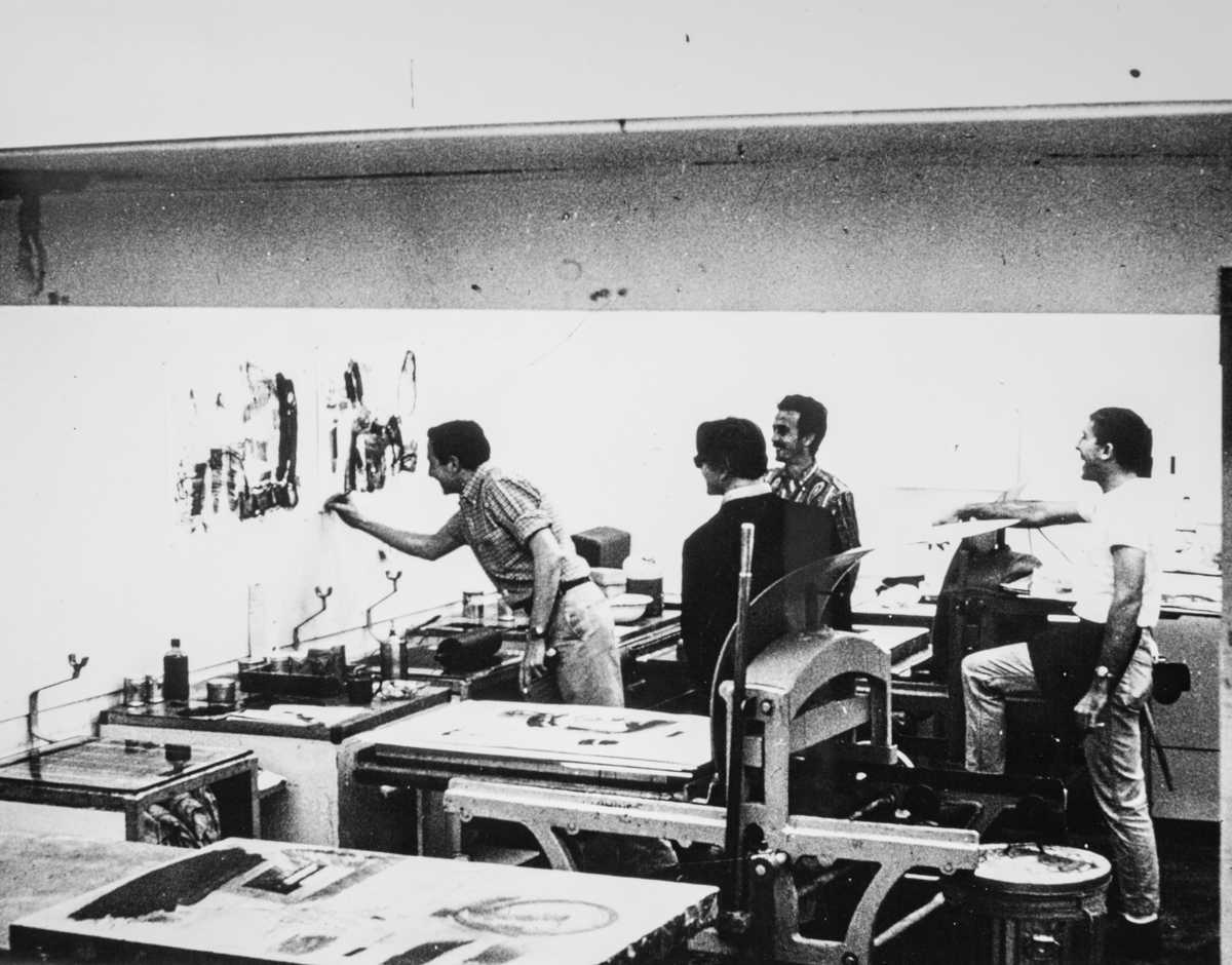 A group of men stand behind some tables and face a wall with one man in front working on an artwork on a wall.